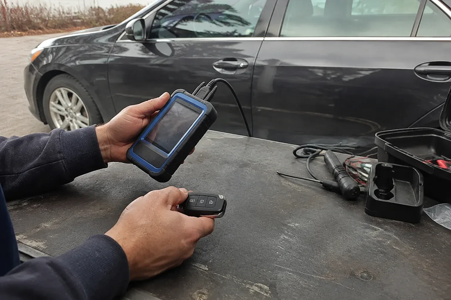Hands holding a blue-edged electronic device and car-key fob at a worktable beside a parked car. — Transponder Key Programming, Georgina